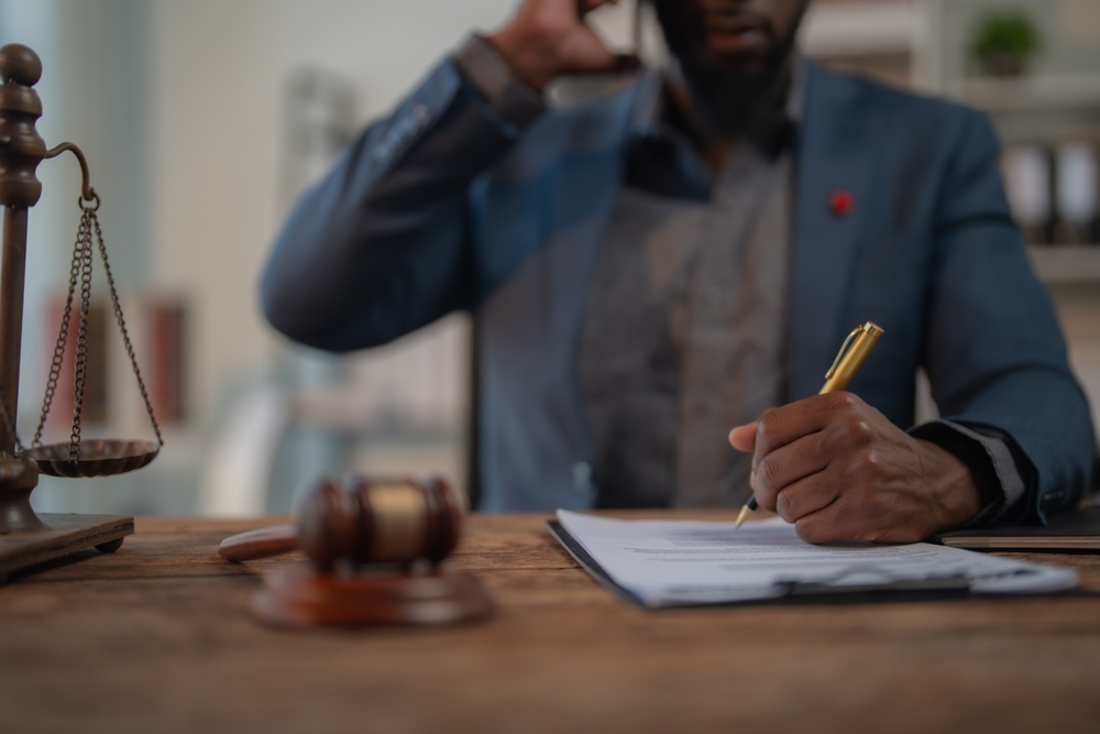 Man on a phone writing on a document at a desk with scales of justice and a gavel